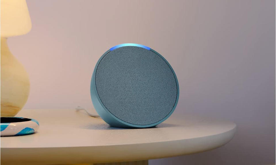 Speaker Amazon Echo Pop berwarna biru terletak di meja malam dengan lampu kuning di dekatnya. 